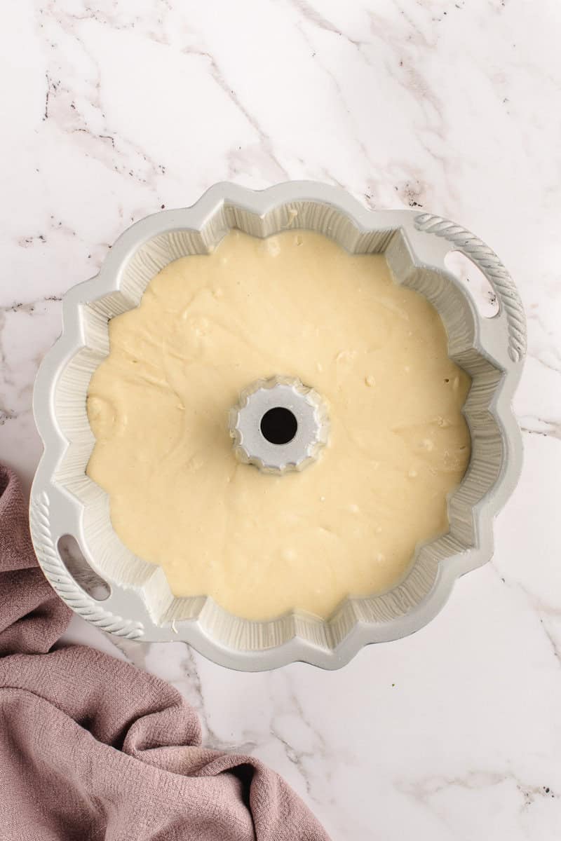 Bundt cake pan filled with cream-colored batter, resting on a marble surface next to a pink cloth.