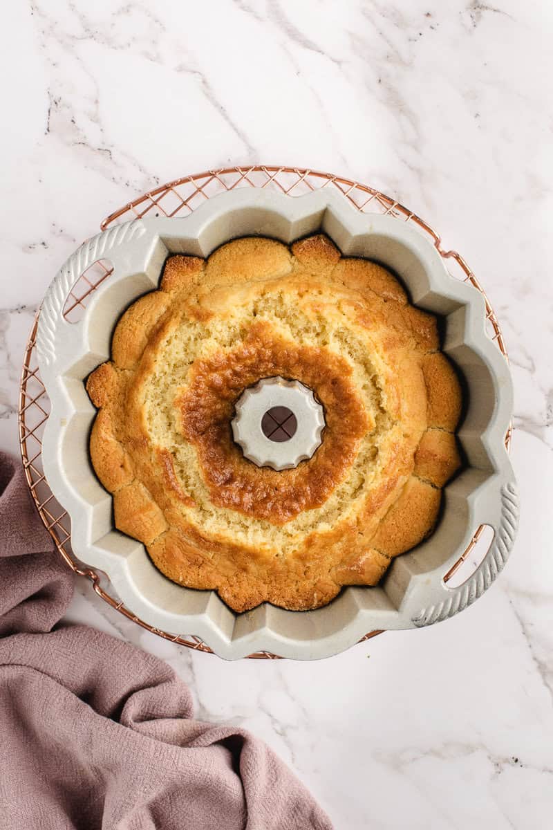 A baked cake sits in a scalloped bundt pan on a marble surface, with a light purple cloth nearby.