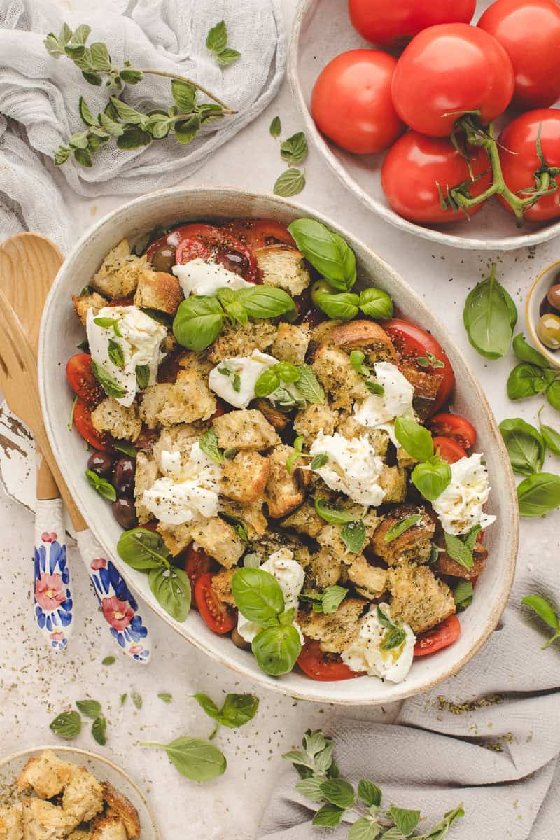A bowl of bread and tomato salad topped with mozzarella and basil leaves on a white table, surrounded by fresh tomatoes and herbs.