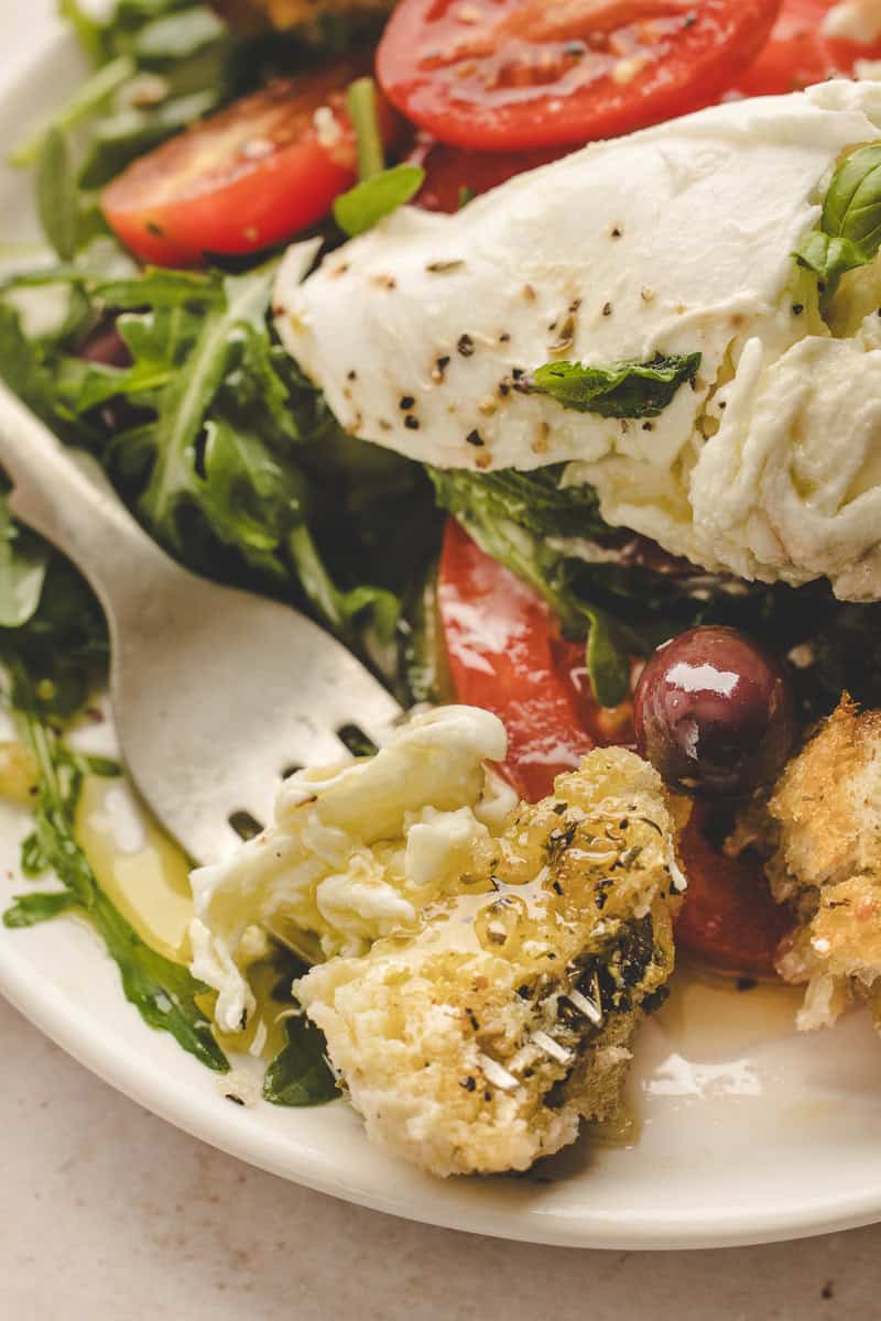 Plate of salad with sliced tomatoes, arugula, mozzarella cheese, olives, croutons, and a fork.