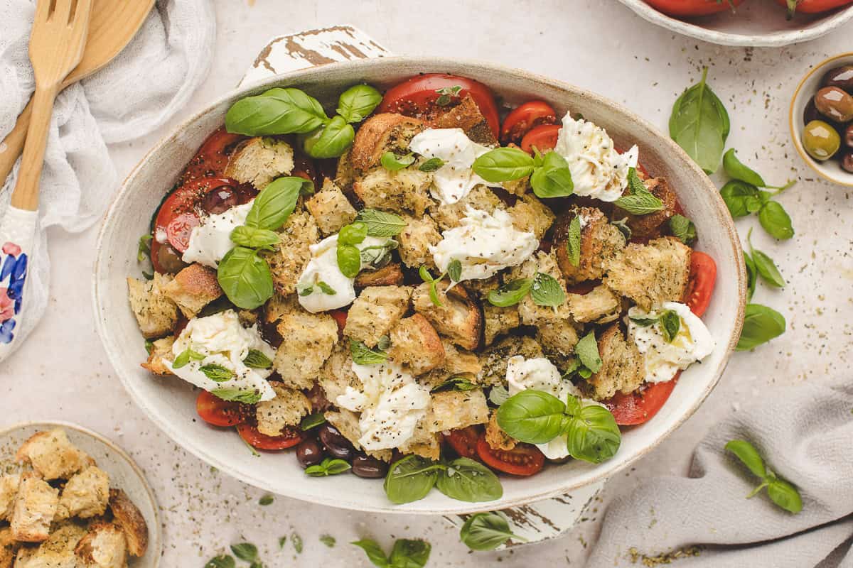 A rustic salad with chunks of bread, mozzarella, tomatoes, olives, and fresh basil in a white dish.