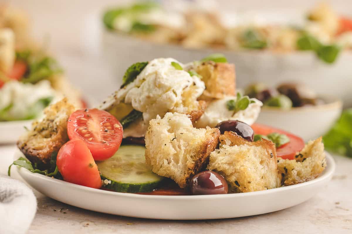 A plate of salad with cherry tomatoes, cucumber slices, olives, croutons, and a dollop of white cheese or cream. Another salad is blurred in the background.