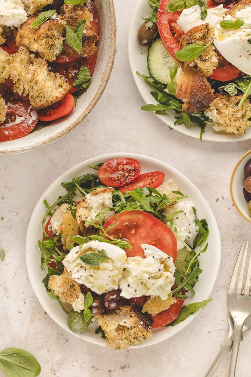 A fresh salad with tomatoes, arugula, cucumbers, croutons, olives, and burrata on a white plate. Fork on the side.