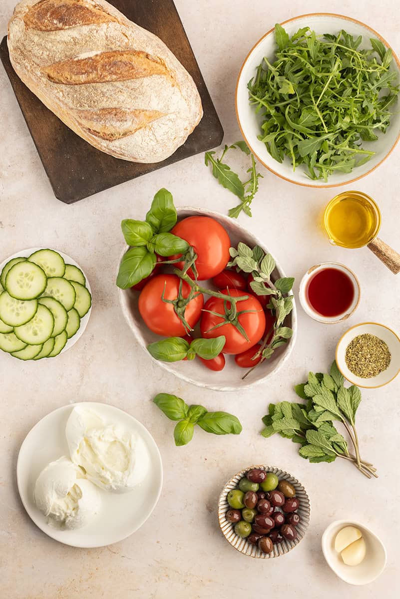 Ingredients for a Mediterranean salad spread on a surface include bread, arugula, tomatoes, herbs, cucumber slices, mozzarella, olives, garlic, and small bowls of oil, vinegar, and seasoning.