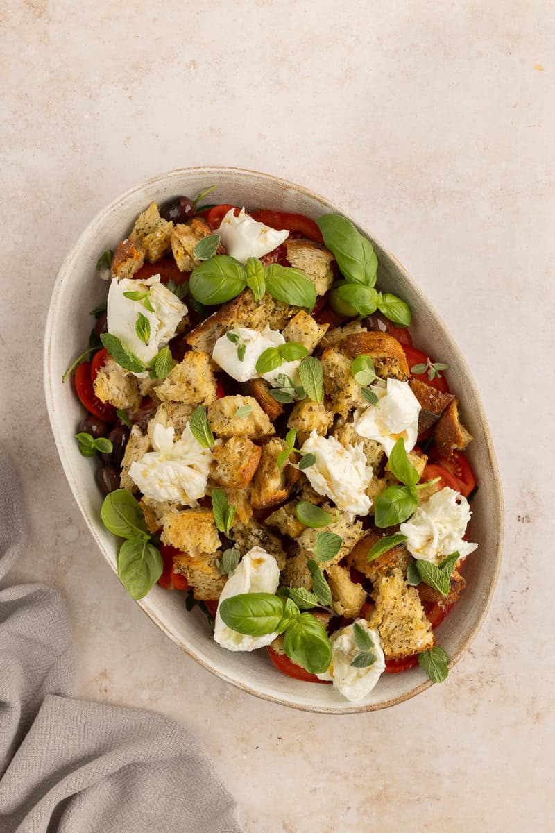 A bowl of panzanella salad with bread pieces, tomatoes, mozzarella, basil, and oregano on a light surface with a gray cloth nearby.