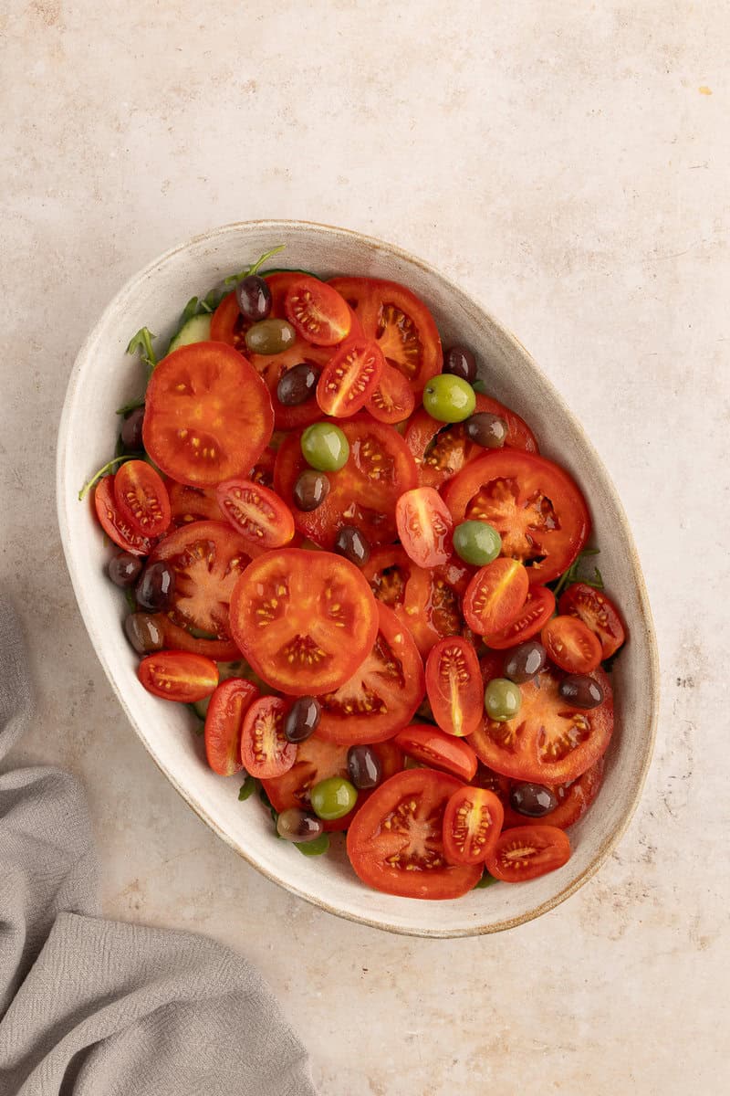 A white oval dish filled with sliced tomatoes, black and green olives on a light textured surface. A gray cloth is partially visible in the bottom left corner.