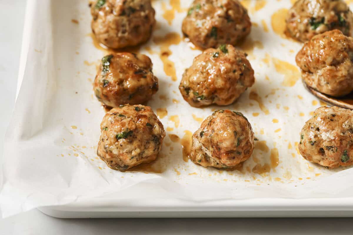 Cooked meatballs with herbs on a parchment-lined baking sheet.