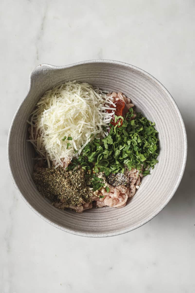 A bowl with minced meat, shredded cheese, chopped herbs, and dried spices on a white surface.
