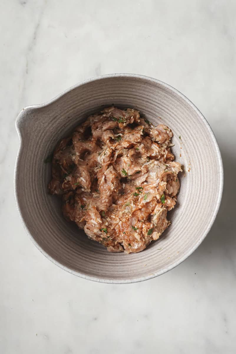 Top-down view of a ceramic bowl containing a mix of minced meat and herbs on a marble surface.