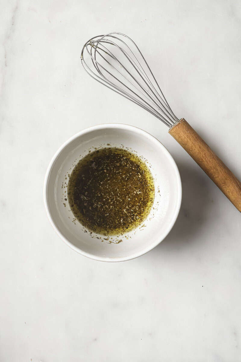 A whisk with a wooden handle lies next to a white bowl containing a mixture of oil and spices on a marble surface.