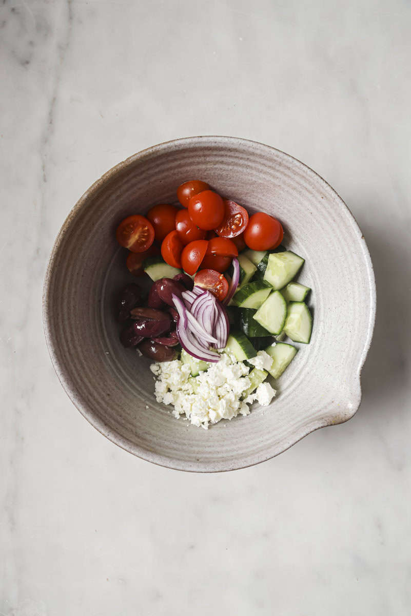 A bowl with cherry tomatoes, sliced radishes, cucumber, olives, red onion, and crumbled feta on a marble surface.