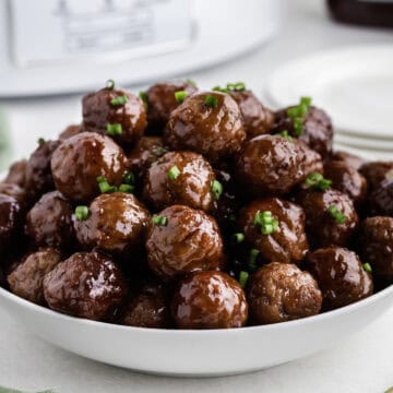 A bowl filled with glossy meatballs, garnished with chopped green herbs, is placed on a table next to a stack of white plates.