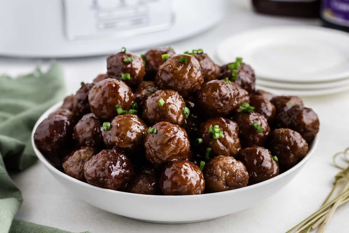A bowl filled with glossy meatballs, garnished with chopped green herbs, is placed on a table next to a stack of white plates.