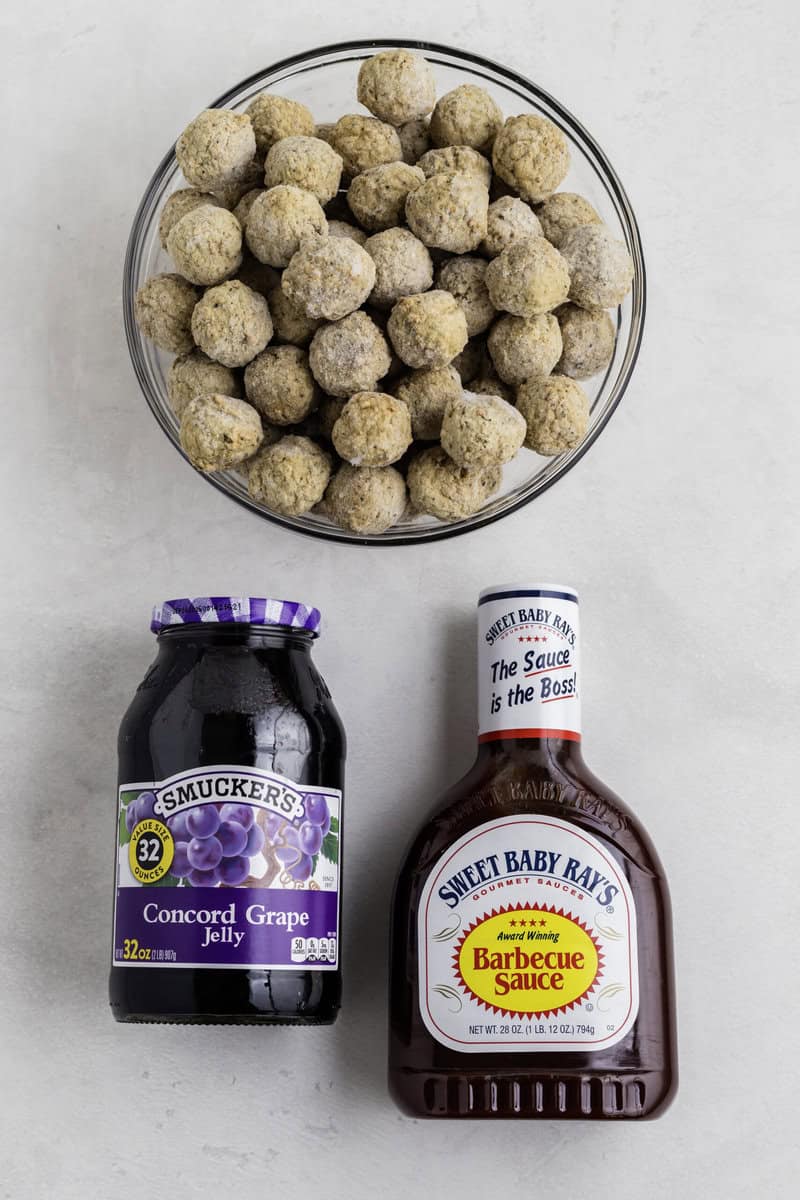 A bowl of frozen meatballs, a jar of Smucker's Concord Grape Jelly, and a bottle of Sweet Baby Ray's Barbecue Sauce are placed on a light surface.