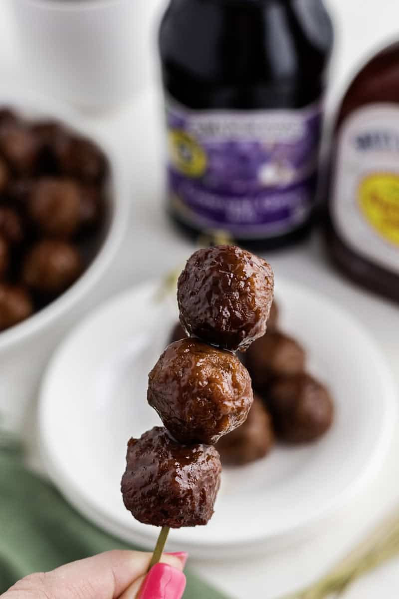 Close-up of three glazed meatballs on a skewer. Blurred background includes a bowl of meatballs, a bottle of sauce, and a plate.