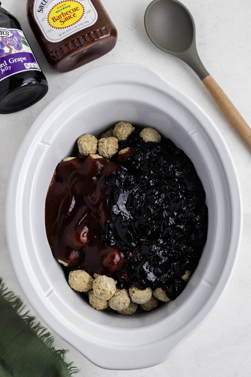 Slow cooker with meatballs covered in barbecue sauce and grape jelly, surrounded by jars and a spoon on a countertop.