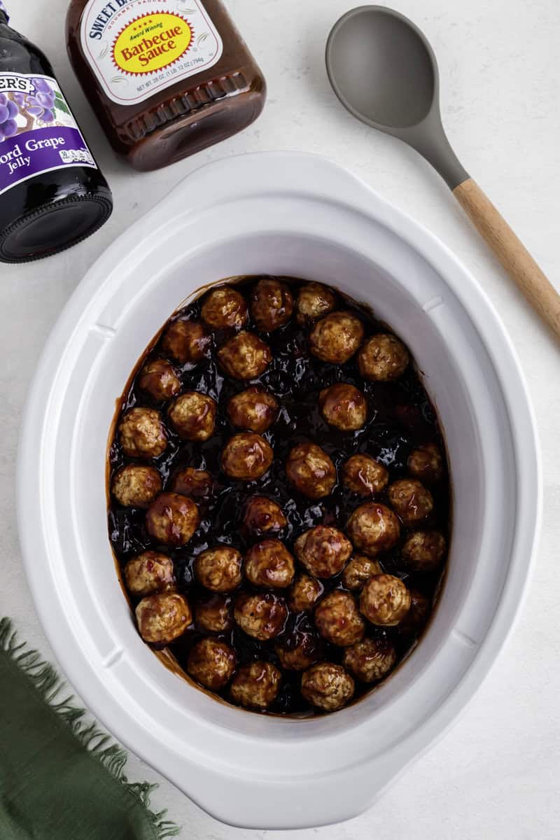 A crockpot filled with cooked meatballs in a sauce, with a jar of grape jelly and a bottle of barbecue sauce nearby. A spoon and green napkin are also visible.