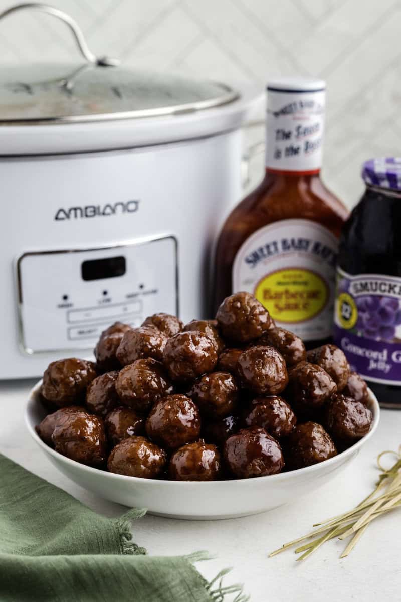 A bowl of glazed meatballs is in front of a slow cooker, with bottles of barbecue sauce and grape jelly nearby.