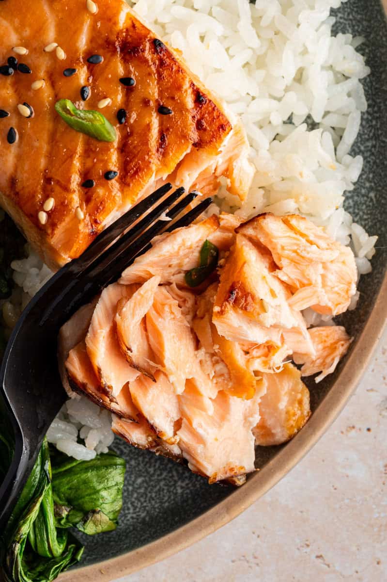 Grilled salmon with sesame seeds, served with rice and greens, partially flaked with a black fork on a plate.