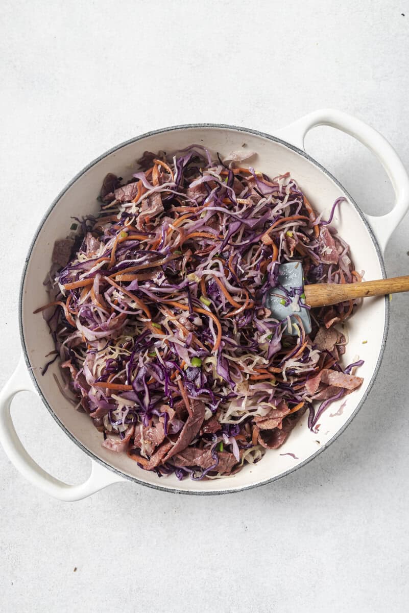 A white pot filled with cooked shredded red cabbage, carrots, and onions, with a wooden spoon resting inside.