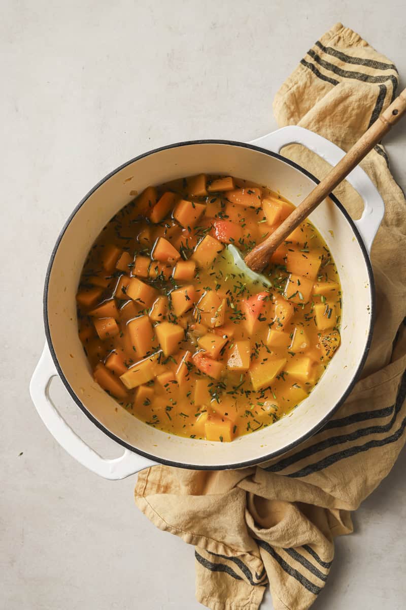 Creamy butternut squash and vegetable soup in a white pot with a wooden spoon, garnished with herbs.