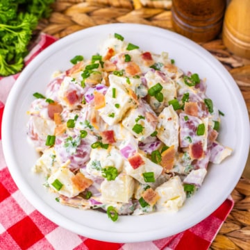 A white plate of creamy potato salad with chopped bacon, green onions, and herbs, set on a red and white checkered napkin beside salt and pepper shakers.