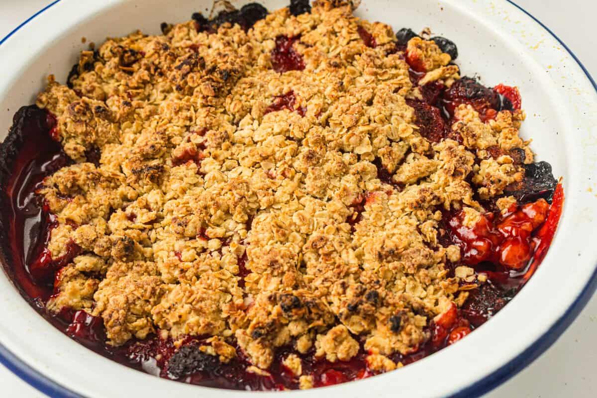 A close-up of a baked berry crumble in a round white dish, showing a golden oat topping over cooked mixed berries.