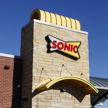 Exterior view of a Sonic Drive-In restaurant showing a yellow and red Sonic logo sign on a brick and stone wall under a clear blue sky, inviting guests to explore their Allergen Menu with gluten free options available.