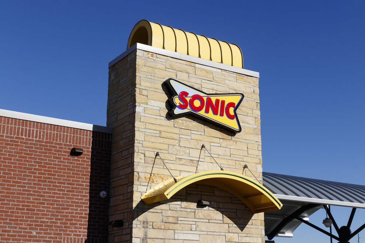 Exterior view of a Sonic Drive-In restaurant showing a yellow and red Sonic logo sign on a brick and stone wall under a clear blue sky, inviting guests to explore their Allergen Menu with gluten free options available.