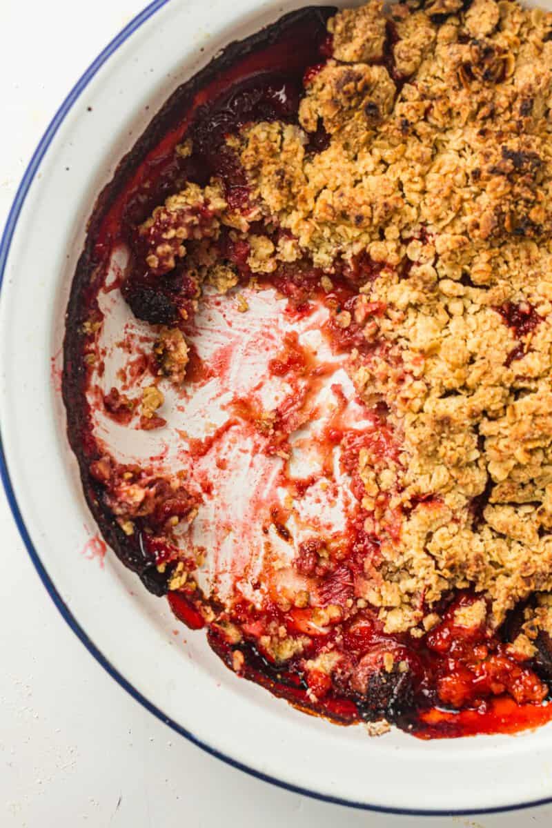 A partially eaten berry crumble in a white dish, showing a mix of golden oat topping and red berry filling.