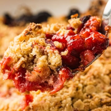 A close-up of a spoonful of fruit crumble dessert, showing a golden oat topping and a red fruit filling.