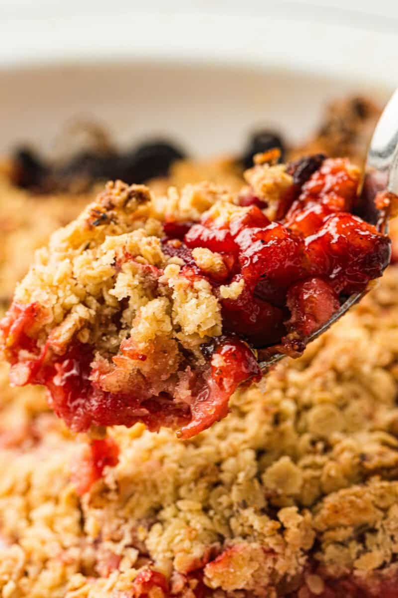 A close-up of a spoonful of fruit crumble dessert, showing a golden oat topping and a red fruit filling.