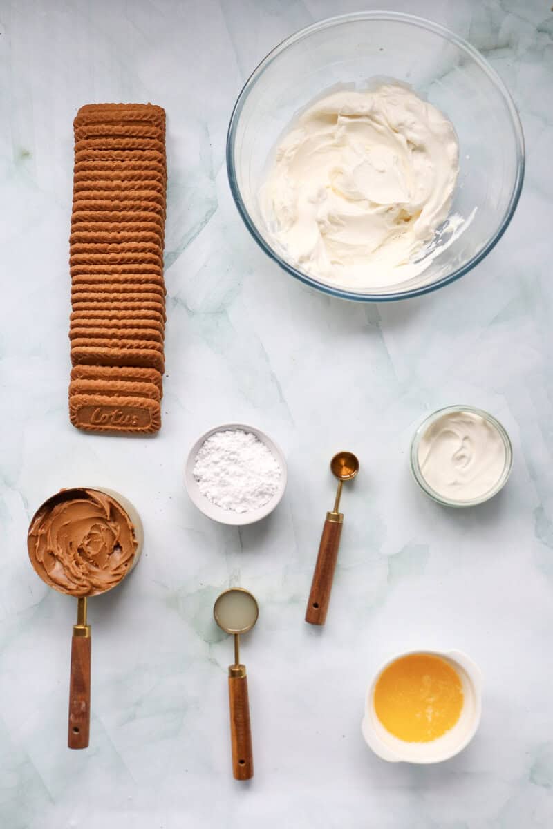 Biscoff cookies and bowls and spoons filled with cookie butter, melted butter, cream cheese, powdered sugar, vanilla extract, and whipping cream on a marble background.