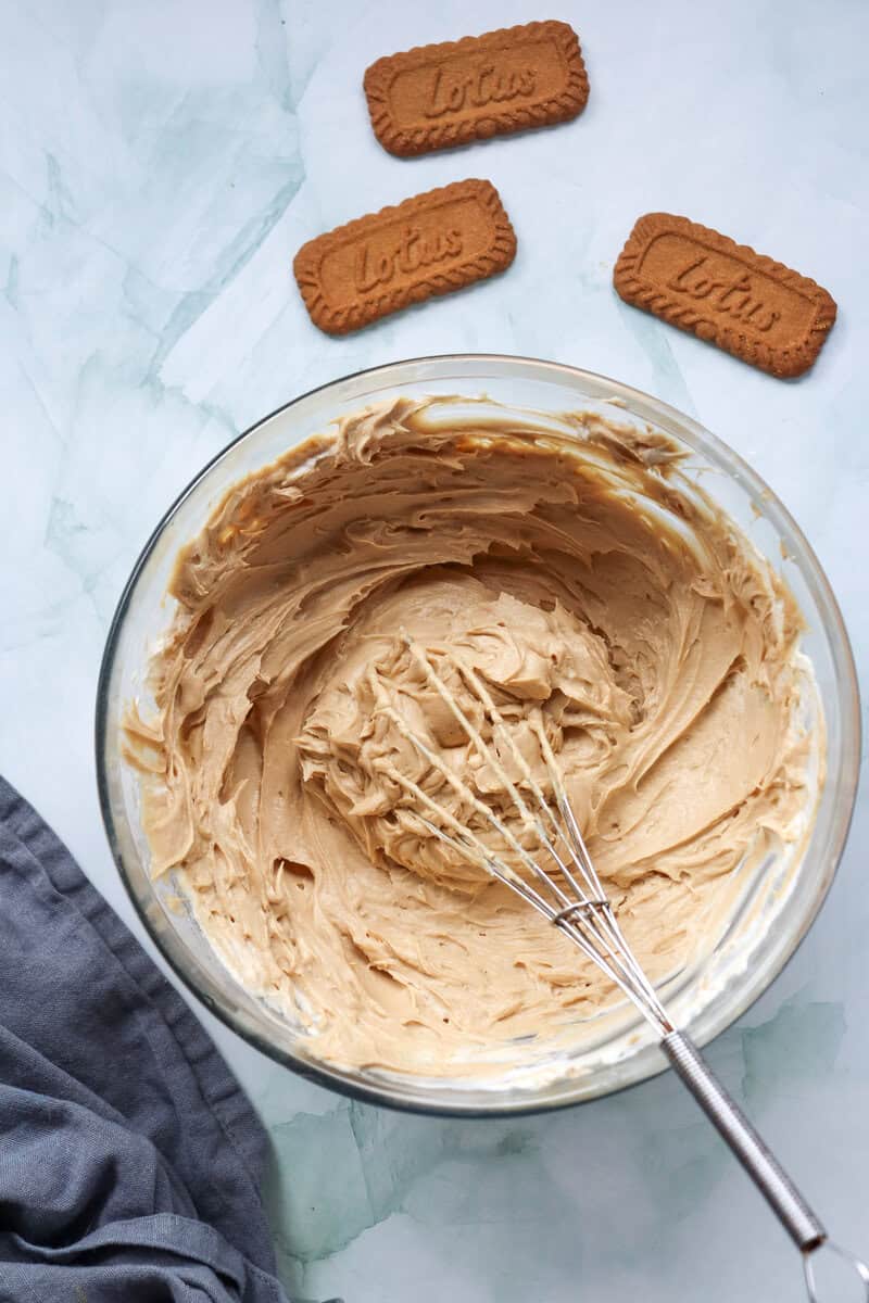 Cream cheese, powdered sugar, vanilla extract, and cookie butter with whipped cream folded in, in a glass mixing bowl with a whisk.