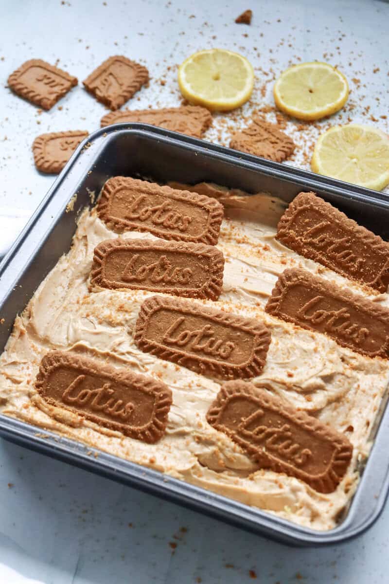 A no bake Biscoff cheesecake topped with Lotus Biscoff cookies with more cookies and lemon slices seen in the background.