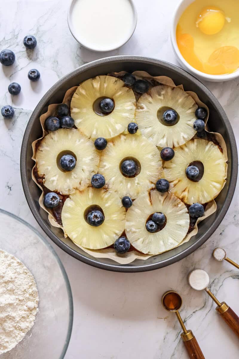 Pineapple rings and blueberries in a cake pan on a white marble countertop.