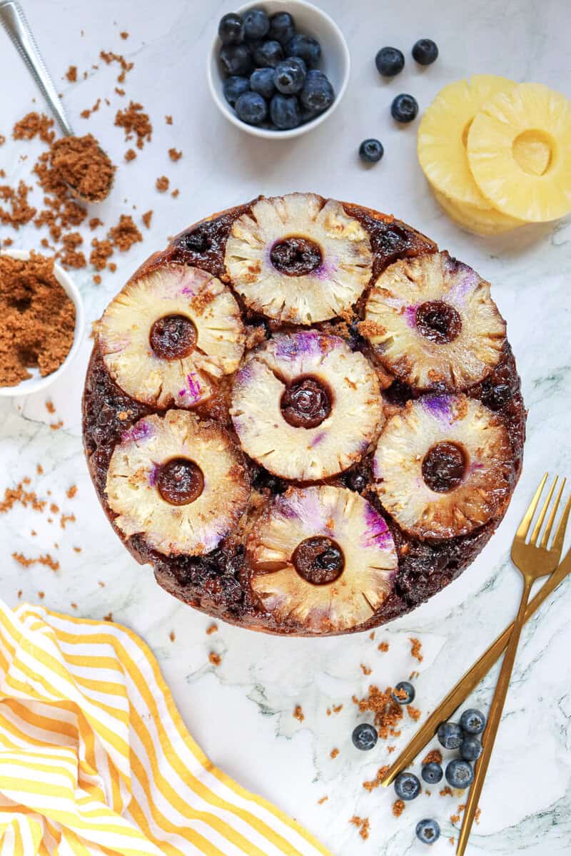 A pineapple blueberry upside down cake on a cake stand with gold forks, a yellow and white striped cloth napkin, blueberries, pineapple rings, and brown sugar scattered around