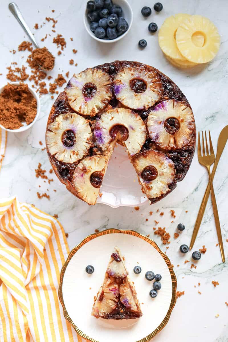 A pineapple upside down cake with blueberries on a marble countertop with a slice removed and placed on a white plate.