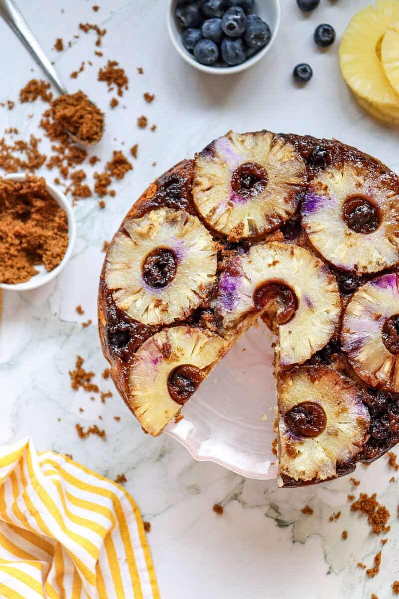 A blueberry pineapple upside down cake with a slice removed on a white background with brown sugar, blueberries, pineapple rings, and a yellow and white linen napkin nearby.