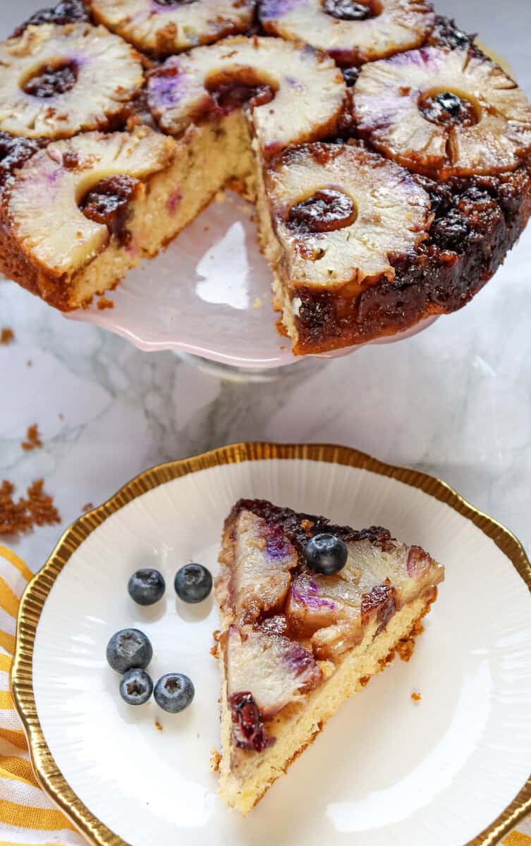 A pineapple and blueberry upside down cake on a white cake stand with a slice removed and placed on a white plate.