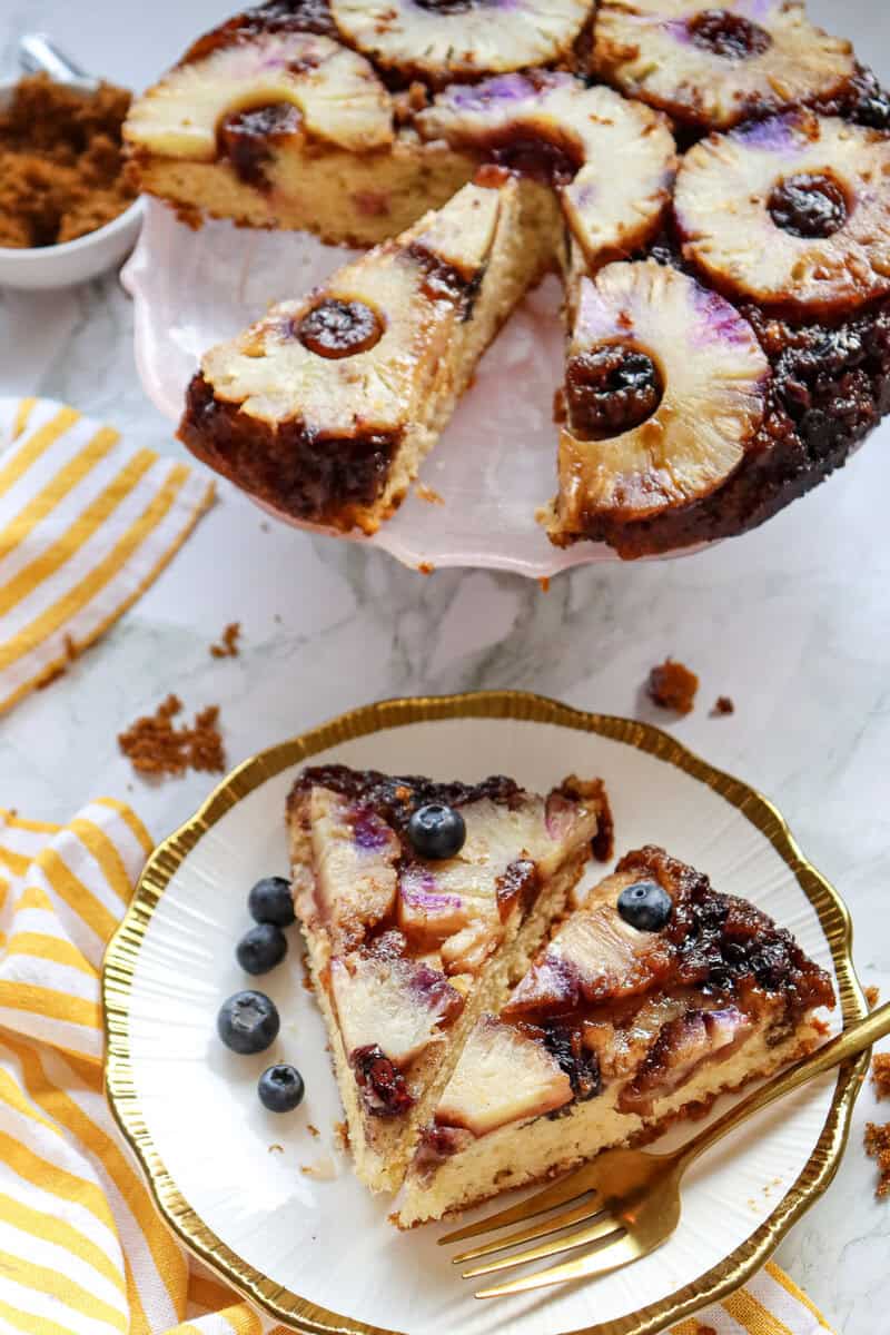 Two slices of pineapple and blueberry cake on a white plate with a gold fork, the whole cake seen in the background.