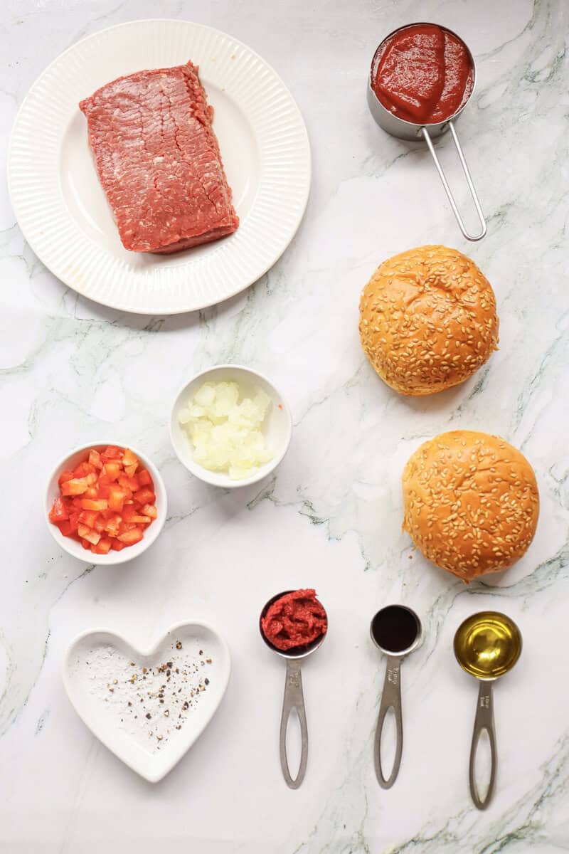 Ground beef, hamburger buns, onions, tomato paste, red bell peppers, and spices on a white marble counter.
