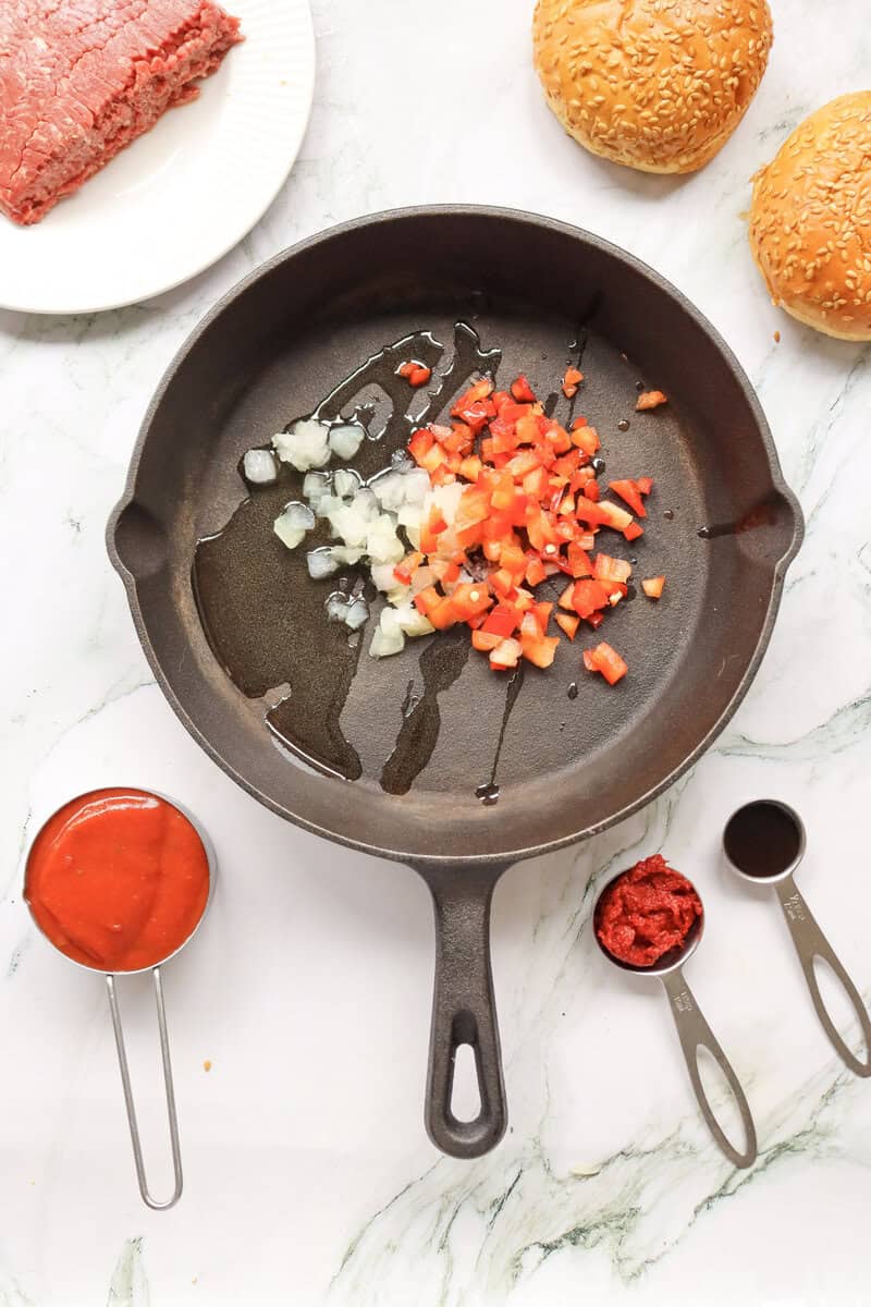 A cast iron skillet with olive oil, onions, and diced red pepper with other ingredients seen nearby.