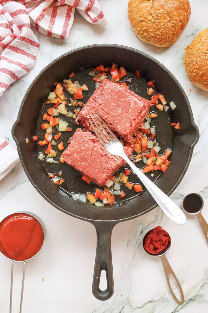 A cast iron skillet with raw ground beef, cooked vegetables, and a silver fork on top.