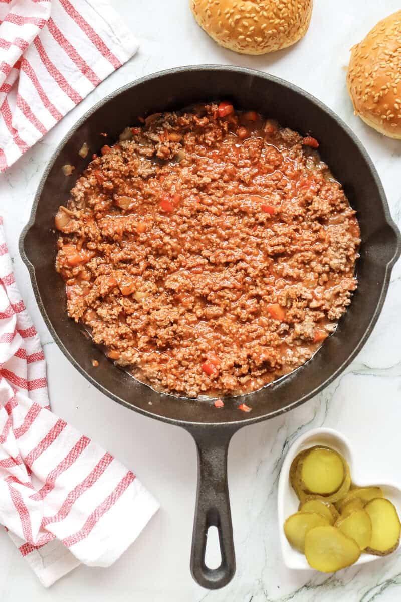 A skillet of homemade Sloppy Joe filling with a heart-shaped bowl of pickles seen nearby.