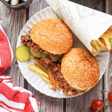 Two homemade Sloppy Joes on a white plate with a paper cone filled with French fries on a wooden background.