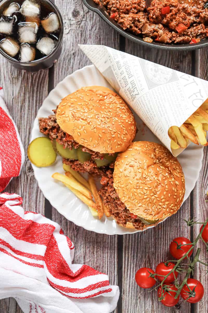 Two homemade Sloppy Joes on a white plate with a paper cone filled with French fries on a wooden background.