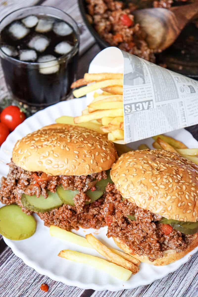Homemade Sloppy Joes on a white plate with pickle slices, french fries, and a glass of soda seen in the background. 