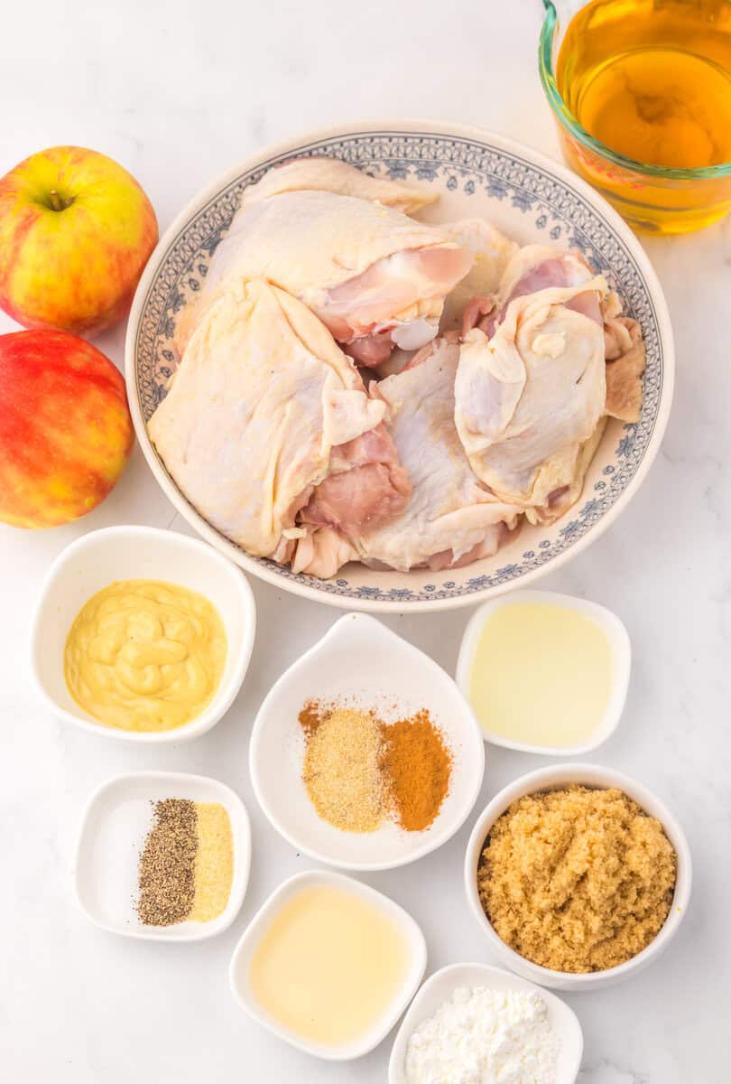 A bowl of raw chicken thighs with seasoning, apples, apple cider, vinegar, dijon, and brown sugar in individual bowls.