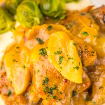 Apple cider chicken with sliced apples and sauce served over mashed potatoes, garnished with chopped herbs, with Brussels sprouts on the side.